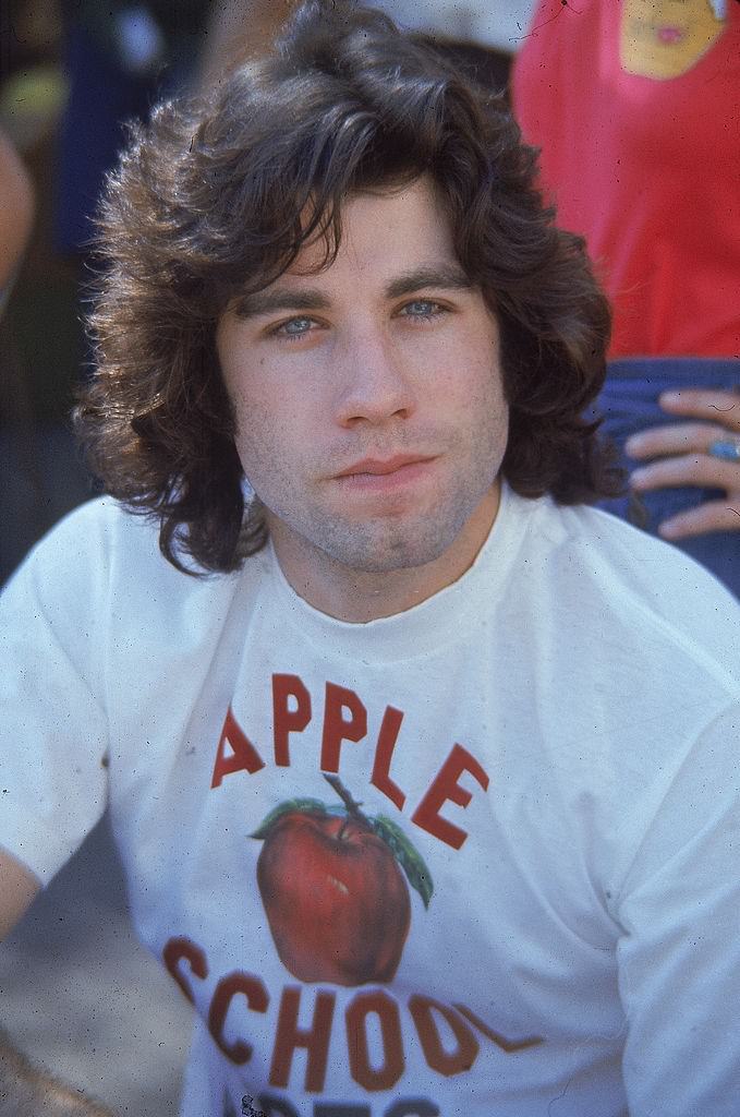 Travolta poses wearing T shirt with the words, 'Apple School,' and an image of an apple emblazoned across the front, 1976