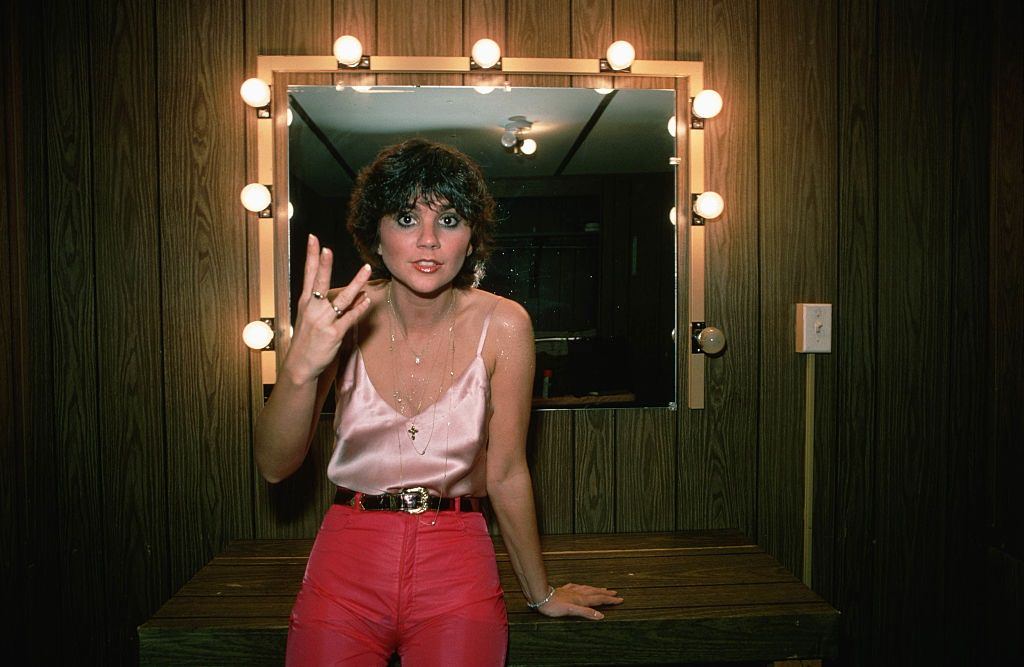Linda Ronstadt standing in front of a mirror with lights around it. She is wearing a pink tank top and hot pink pants with a black belt, 1978