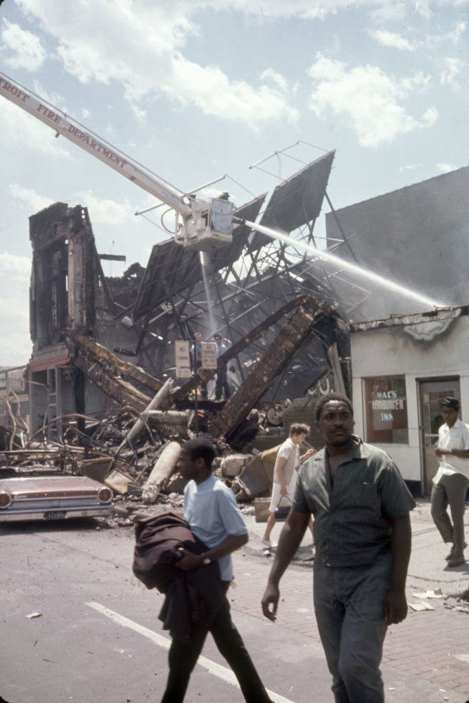 View of a firefighters as they battle a blaze as pedestrians walk beneath them in the wake of ongoing riots, Detroit, Michigan, 1967