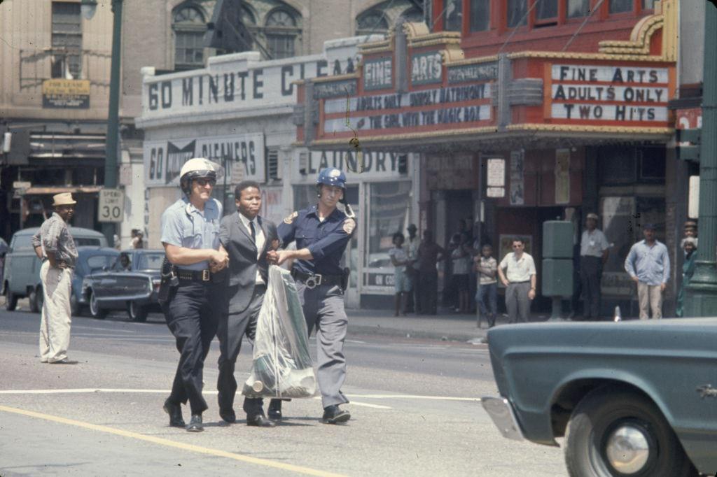 A pair of police officers arrest a young man, dressed in a suit, and escort him down the middle of Woodward Avenue, Detroit, 1967