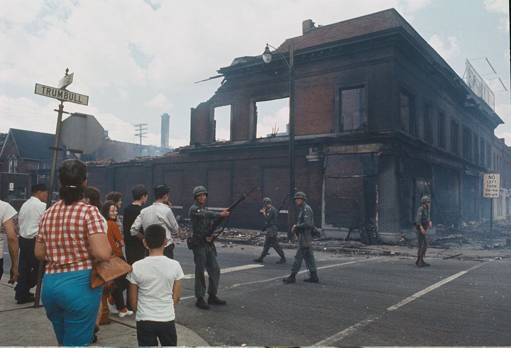 A mellékállók megfigyelik a nemzetőrségeket, akik puskafegyverekkel vannak felfegyverkezve és őrzik az utcai sarkot egy kiégett téglaépület mellett Detroit nyugati oldalán, 1967