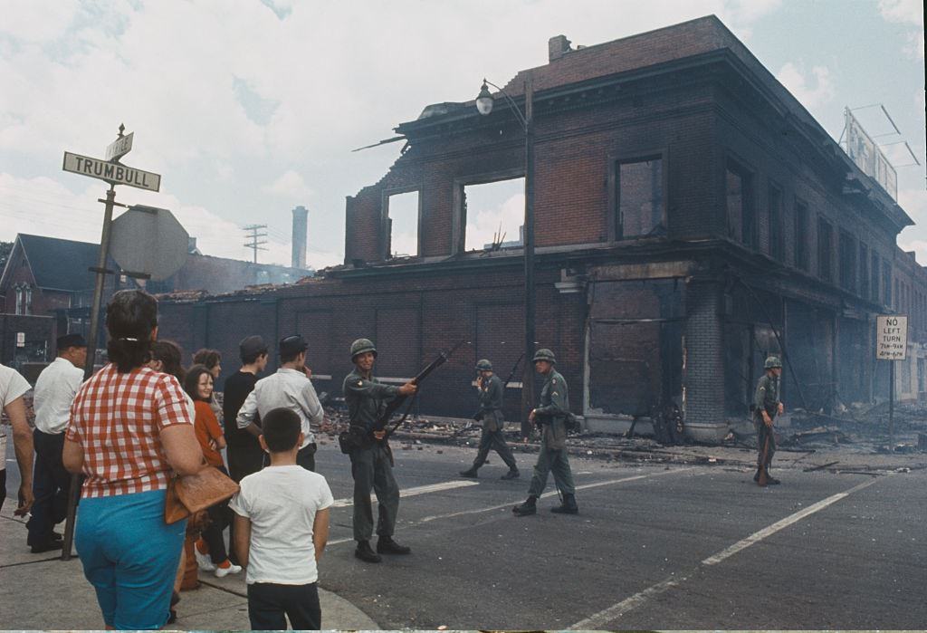 Bystanders observe National Guardsmen, armed with rifles standing guard and patrolling a street corner beside a burnt out brick building on the West Side of Detroit, 1967