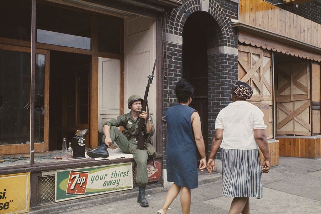 A National Guardsman armed with a rifle and bayonet rests in the window of a wrecked shop front in a shopping street in Detroit, 1967
