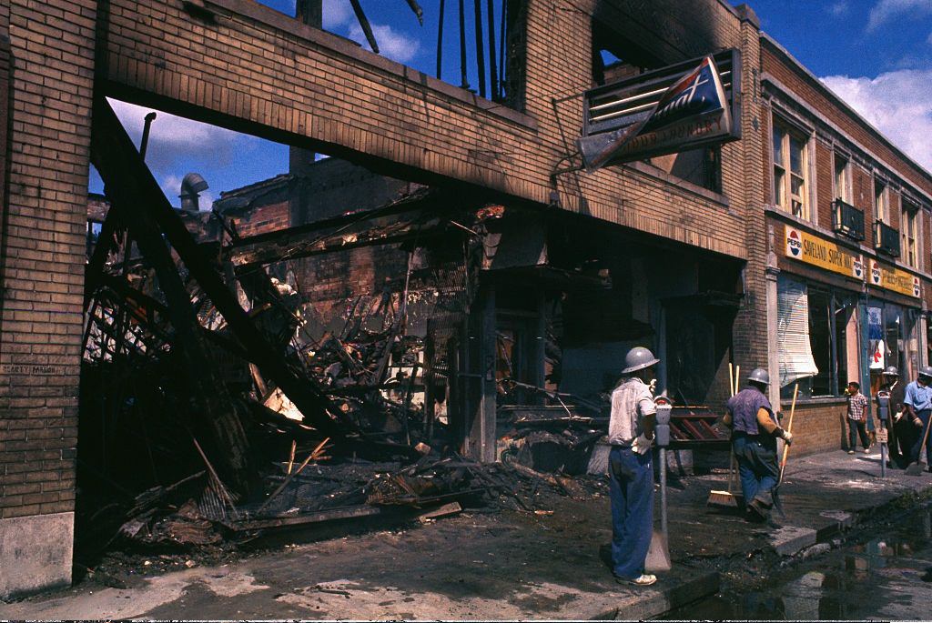 Negro workers shovel and sweep away the sidewalk debris outside the gutted remains of a building, in Detroit, 1967