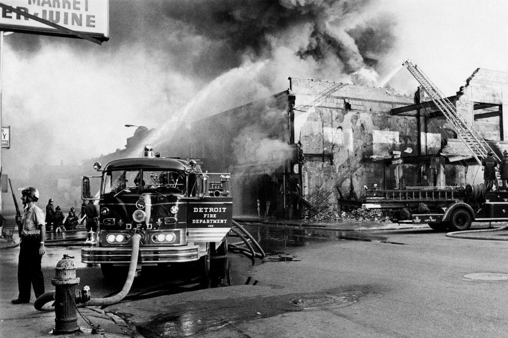 A policeman stands guard in a Detroit street on July 25, 1967 as firefighters try to extinguish a burning building during riots that erupted in Detroit