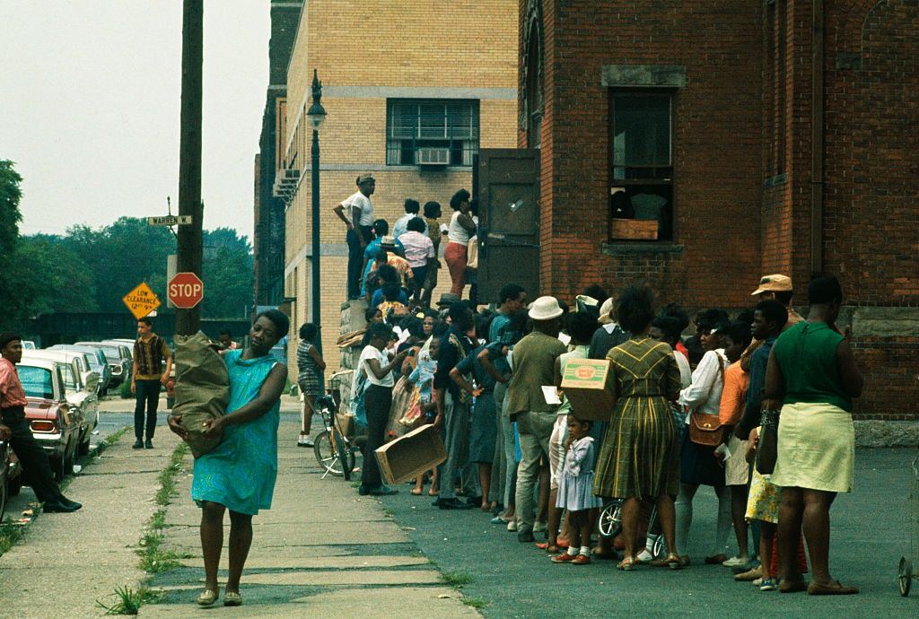 Residents of this riot-torn area, some holding empty cardboard boxes, line up outside building where food and clothing is being distributed, 1967