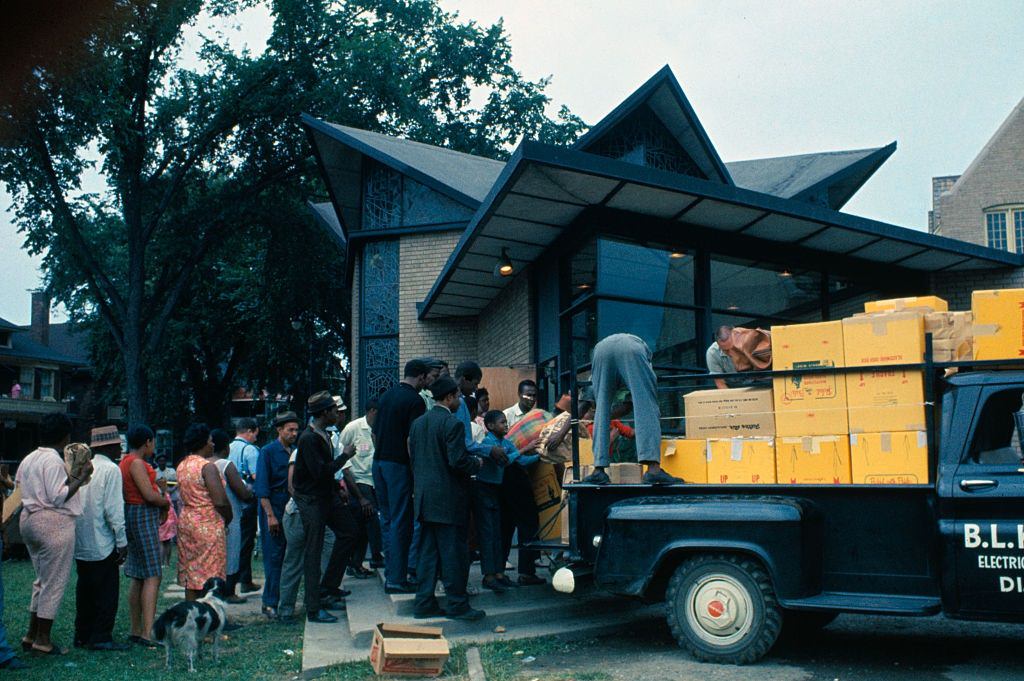 Residents of this riot-torn area, some holding empty cardboard boxes, line up outside building where food and clothing is being distributed, 1967