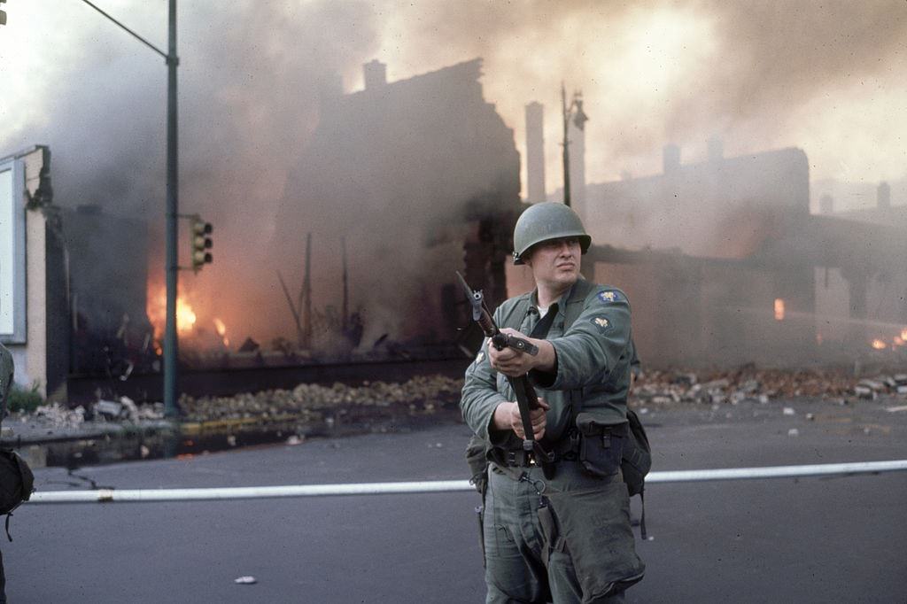 An armed National Guardsman holds is bayonet at the ready as he patrols against a backdrop of buildings that burn in the wake of ongoing riots, Detroit, 1967