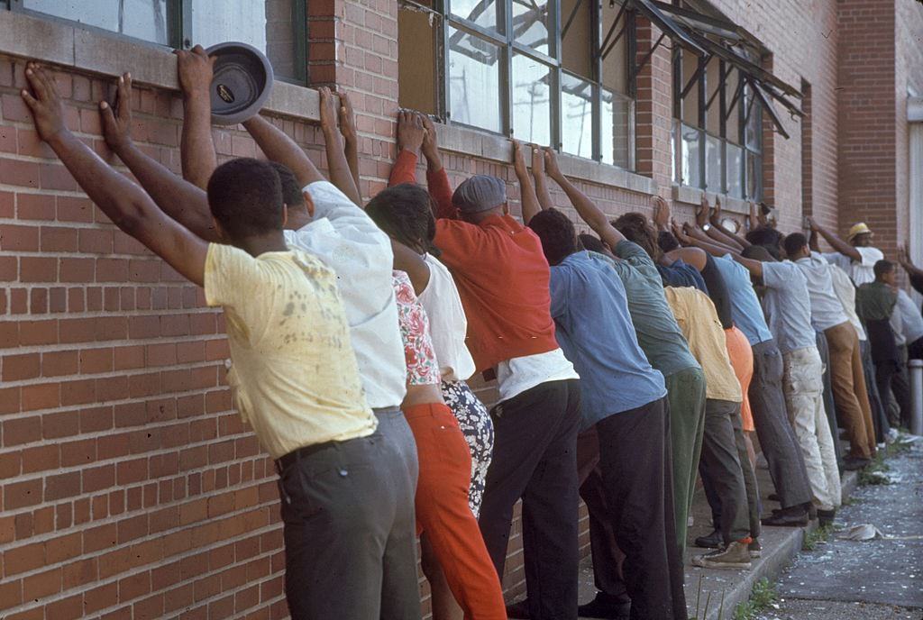 View of male and female African-American suspects lined up, with their hands against a brick wall as they wait to be arrested in the wake of ongoing riots, 1967