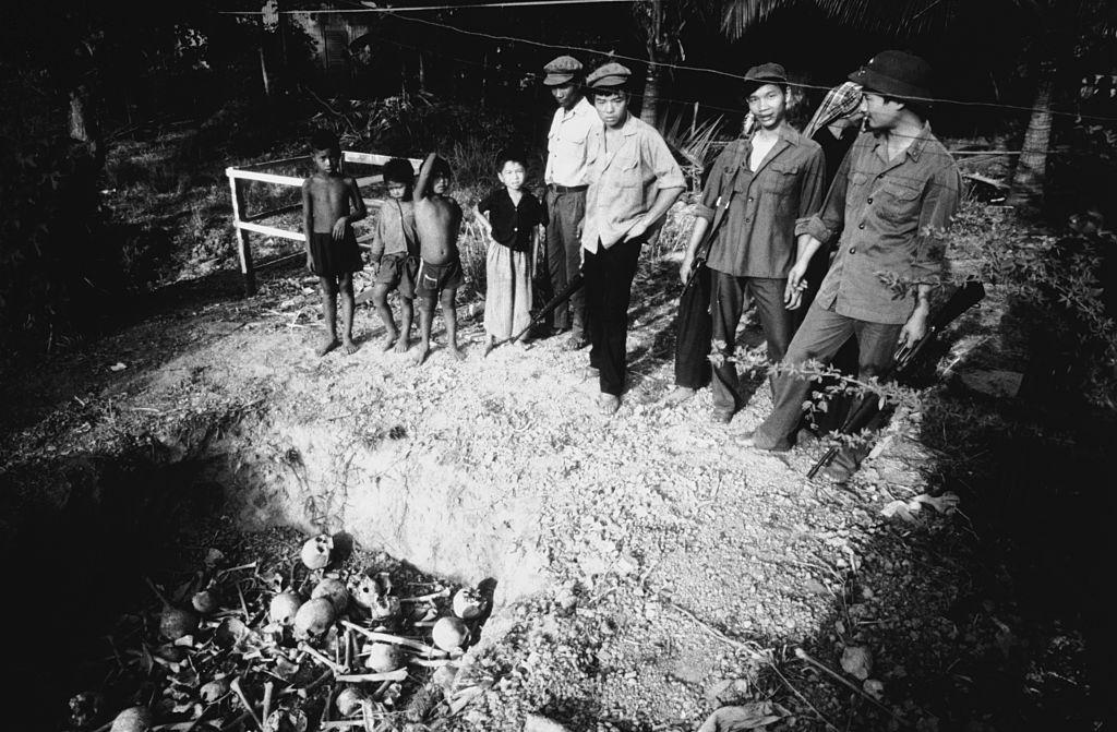 Vietnamese soldiers and a group of children witness the unearthing of a mass grave, 1980