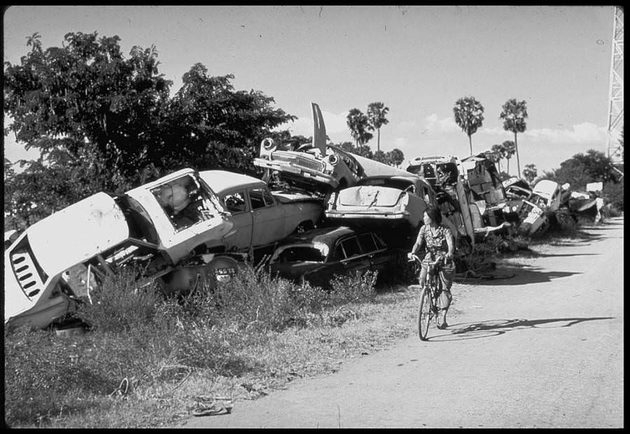 A woman rides a bicycle by a stack of destroyed cars, cast aside by the Khmer Rouge as of symbol of the bourgeoisie, 1979