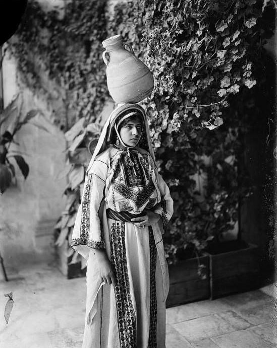 A woman poses with a jug on her head.Ramallah, Circa 1898-1914