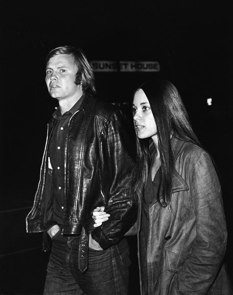 Marcheline Bertrand with Jon Voight walk hand-on-arm along a street at night in front of a sign which reads 'Sunset House,' 1970s.