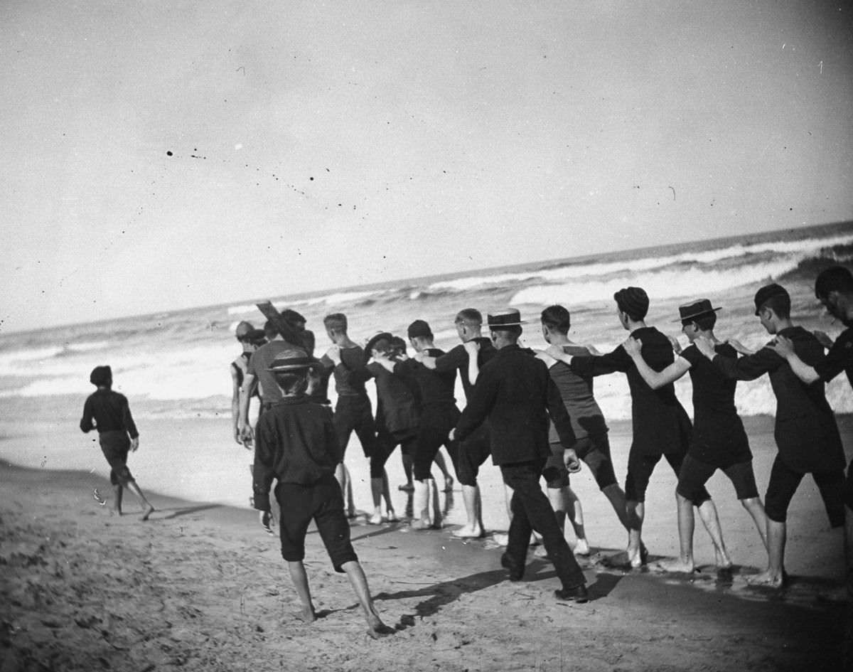 A group of young male bathers walk single file along the beach.Aug. 23, 1886
