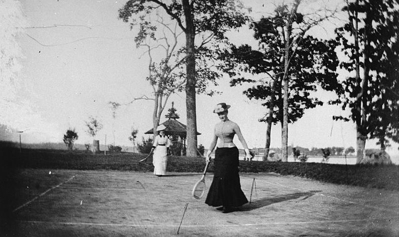 Two well-dressed women playing lawn tennis next to a lake, New York, 1880s