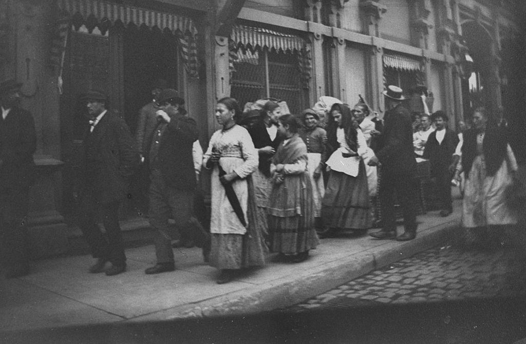 Italian immigrants walking down a narrow sidewalk near the Fulton Ferry, New York, 1880s