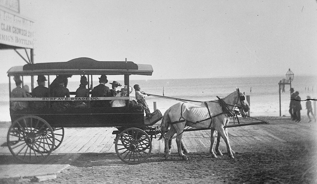 A young black boy pulling the reins of a horse drawn coach owned by the Surf Ave. Stage Co., alongside the boardwalk at Coney Island, 1884