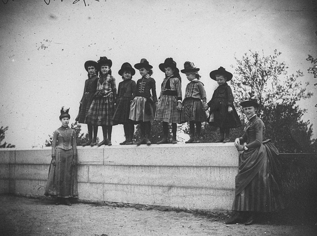 Seven young girls incl. Isabel and Nellie Harter, Nellie and Edie Dwight, Zelma Levison and Edie Swan standing on a wall with two women, New York, 1886