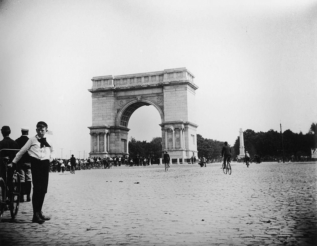 A boy leans against a bicycle in the foreground as people in the background ride their bikes through the arch at Prospect Park during a bicycle parade, Brooklyn, New York, June 15, 1895.