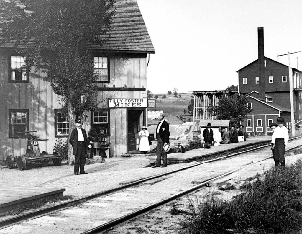 People hanging around outside railroad station, which has signs on it saying TILLY FOSTER MINES, New York, 1889