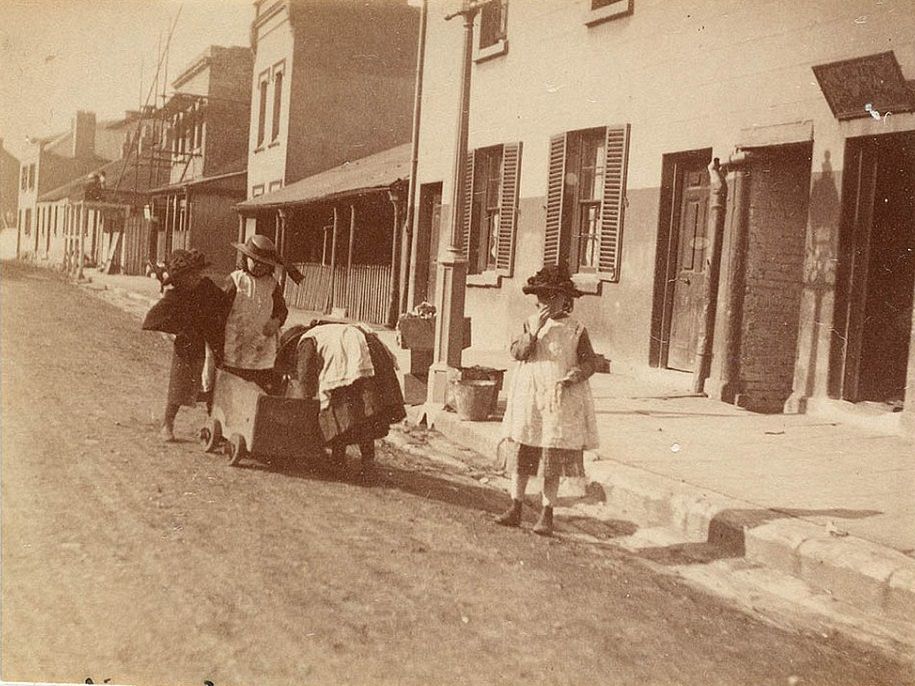 Four children playing in the street in Sydney with a little wooden cart, wearing dresses and hats with ribbons, circa 1885-1890.