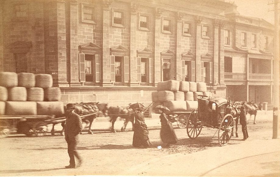 Horses and carts stacked with what appear to be bales of wool being transported through the city, while women in long dresses shaded with umbrellas cross the street, circa 1885-1890.