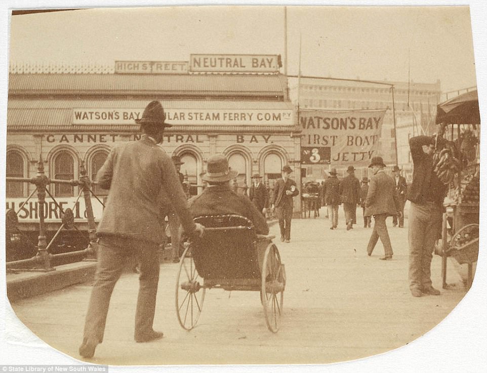 A men pushing another person along in a wheelchair at Circular Quay, circa 1885-1890. Signs advertising the ferry to Watsons Bay are visible, as vendors sell bananas and fruit from stalls on the side of the street .