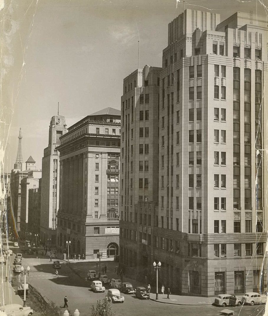 A street in the city in 1947 shows the Rural and Commonwealth Banks on one side, while automobiles driving past and pedestrians crossing the street and on the footpaths