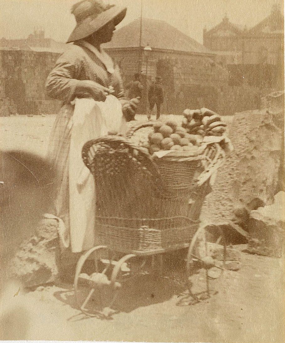 A woman wearing a bonnet and apron is pictured selling fruit from a small wicker barrow with large metal wheels in Sydney circa 1885-1890