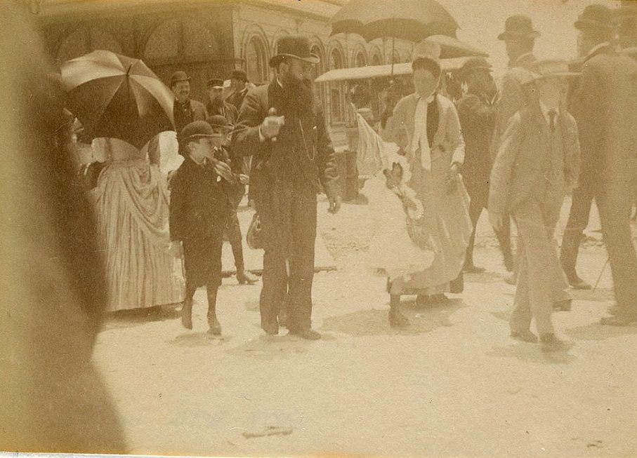 A group of people is seen walking through the streets of Sydney circa 1885-1890, with the men and boys wearing top hats and suits and the women in long dresses carrying umbrellas