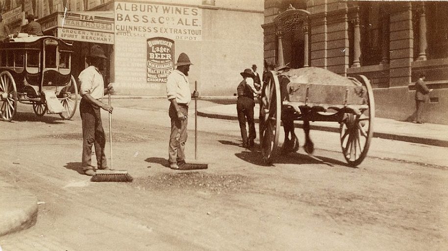 Two men sweeping the dirt street, while horse and carriages and horse and carts trundle past, circa 1885-1890.