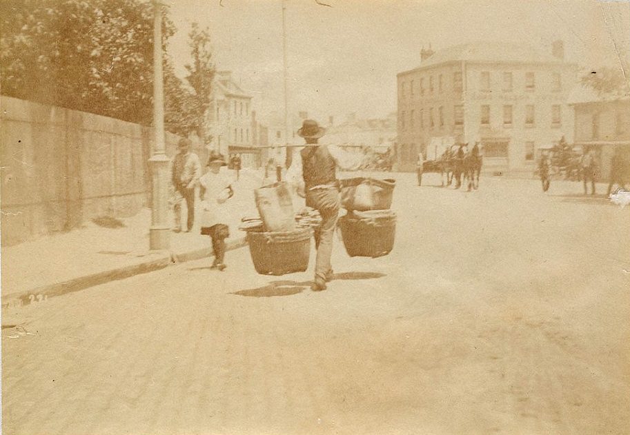 A street vendor transporting goods in baskets stacked on top of each other hanging off either end of a pole balanced on the man's shoulder, taken circa 1885-1890