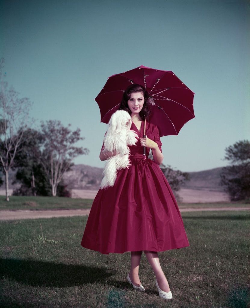 Pier Angeli wearing a red dress and matching parasol, and holding a small white dog, circa 1955