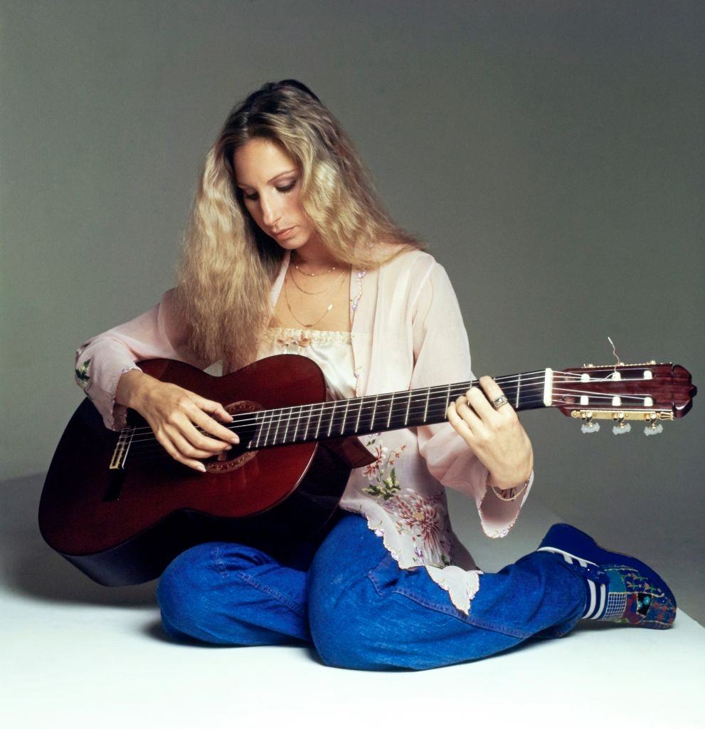 Barbra Streisand wearing blue jeans and a Balinese jacket and playing a guitar, 1975