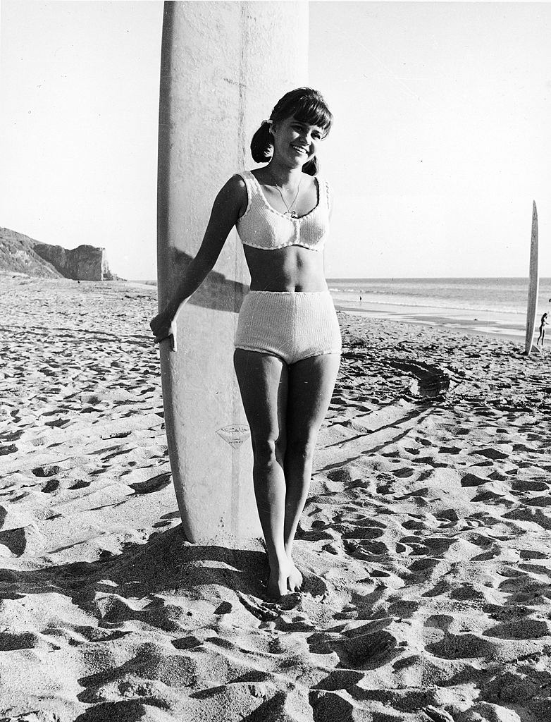 Sally Field in a swimsuit with a surfboard on the beach, circa 1965