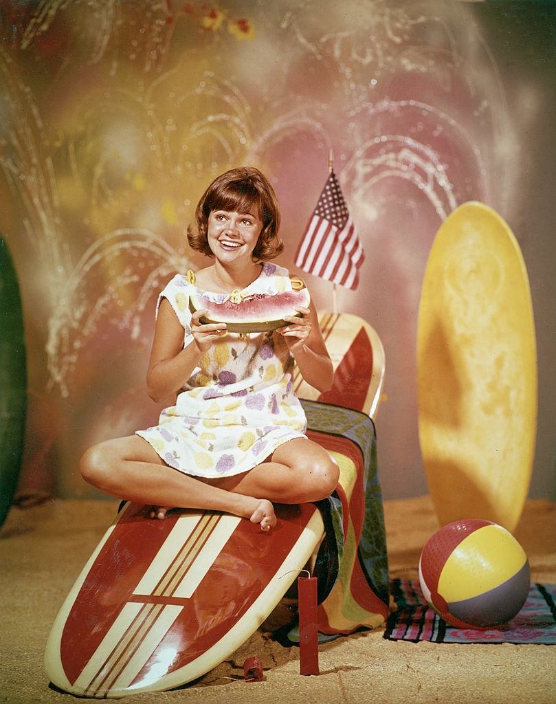 Sally Field sitting on a wooden surfboard with an American flag and eating a watermelon, for the television series, 'Gidget,' 1965