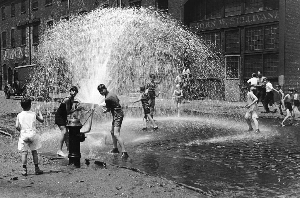 A group of children in shorts play in an open fire hydrant on a cobblestone street, New York City.