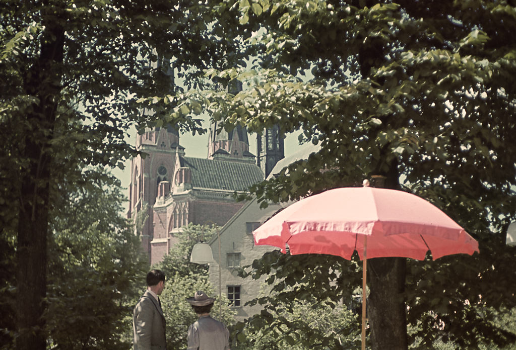 A man and a woman, a pink parasol and Uppsala Cathedral. The image is taken from the garden of the Skytteanum building, partly viewed in the image, 1943