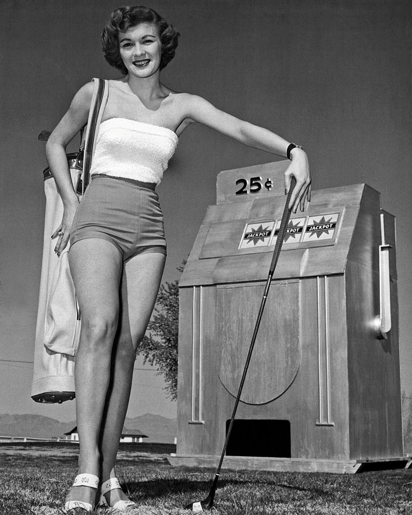 Woman golfer in short shorts stands next to a model of a slot machine on the golf course, Las Vegas, 1956.
