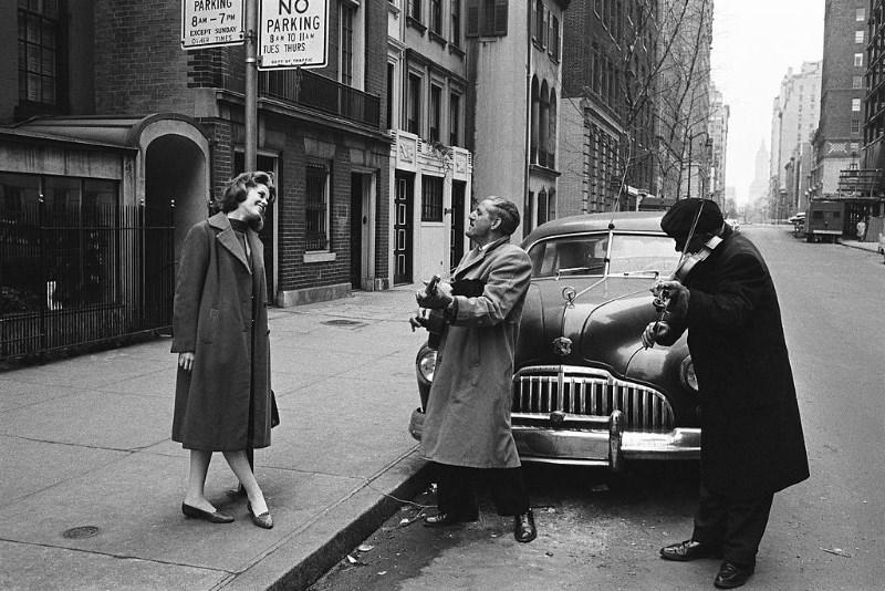 Jane Fonda, leaning against a pole, smiling while listening to the street musicians playing for her.