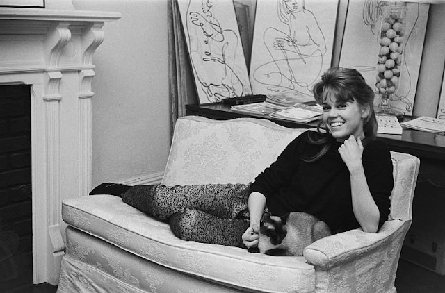 Jane Fonda smiling while lying on the sofa with her Siamese cat at her home.