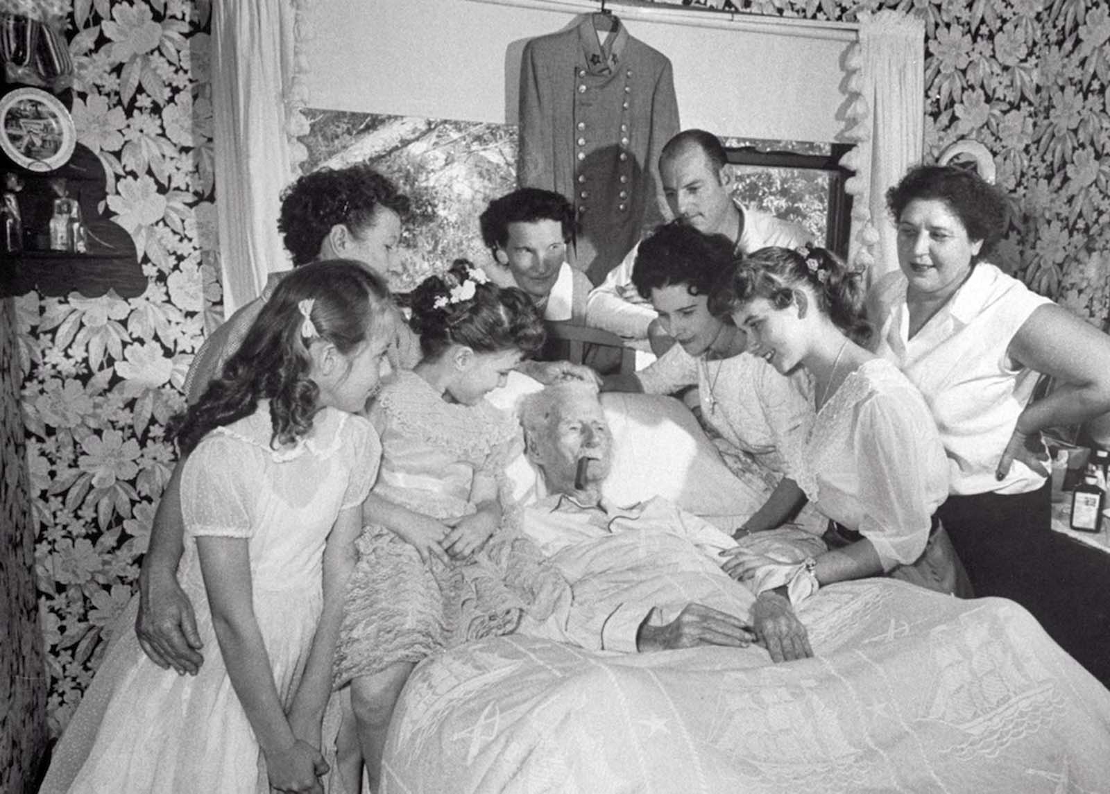 Walter Williams lying in bed with cigar, surrounded by family and friends. 1959.