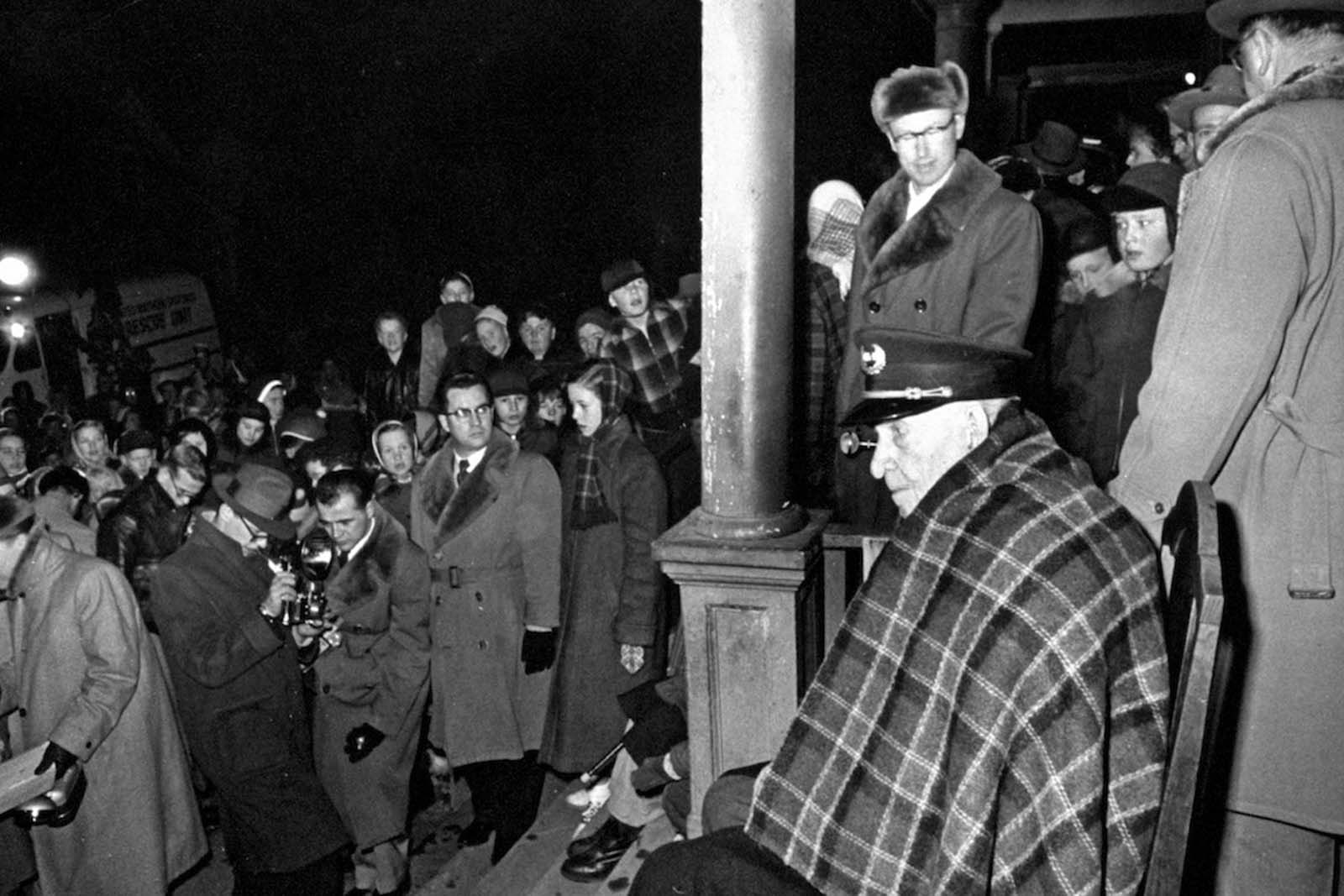 107 years old last remaining Grand Army of the Republic (GAR) Civil War Veteran Albert Woolson (seated) sitting in the front porch wearing a military hat and blanket while people and photographers are passing by. 1954.