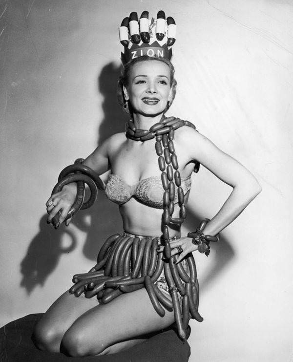 Actor Geene Courtney models a scarf, skirt, bracelets, and a crown made from hot dogs, frankfurters, and kielbasa in her role as Queen of National Hot Dog Week, as selected by the Zion Meat Products Company, 1955