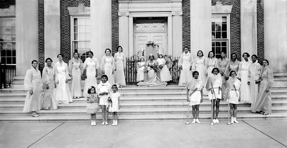 Group of women standing on exterior steps wearing formal dresses. Two groups of three girls stand at the front.