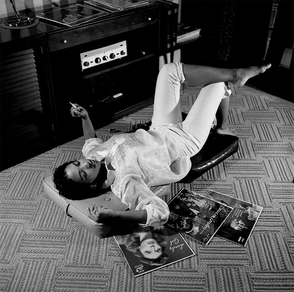 A woman lying down on the floor, smoking cigarette, surrounded by record albums, including “Lonely Girl” by Julie London, released 1956.