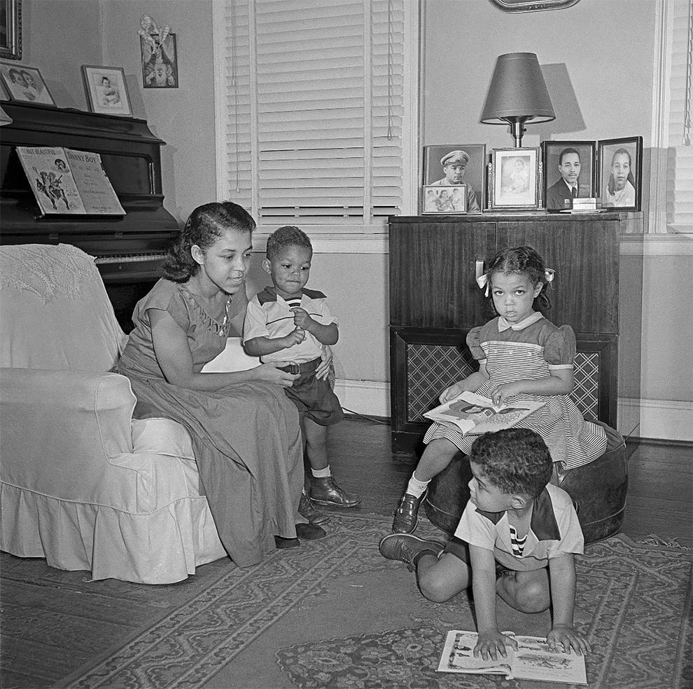 Mother sitting on chair with three children around her, piano to left.