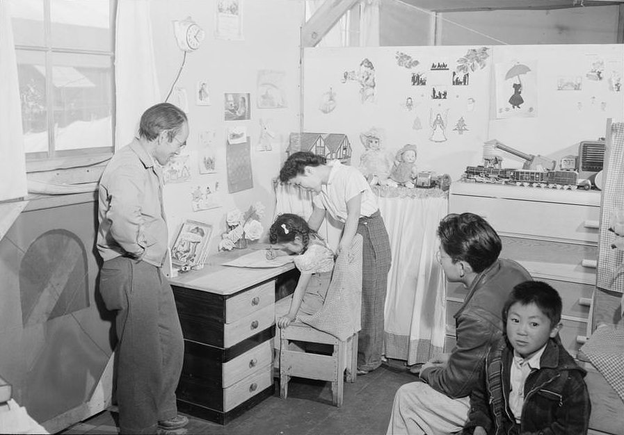 Toyo Miyatake, full-length portrait, standing in his children’s bedroom looking at his young daughter seated at a desk, drawing, while her mother stands behind her.