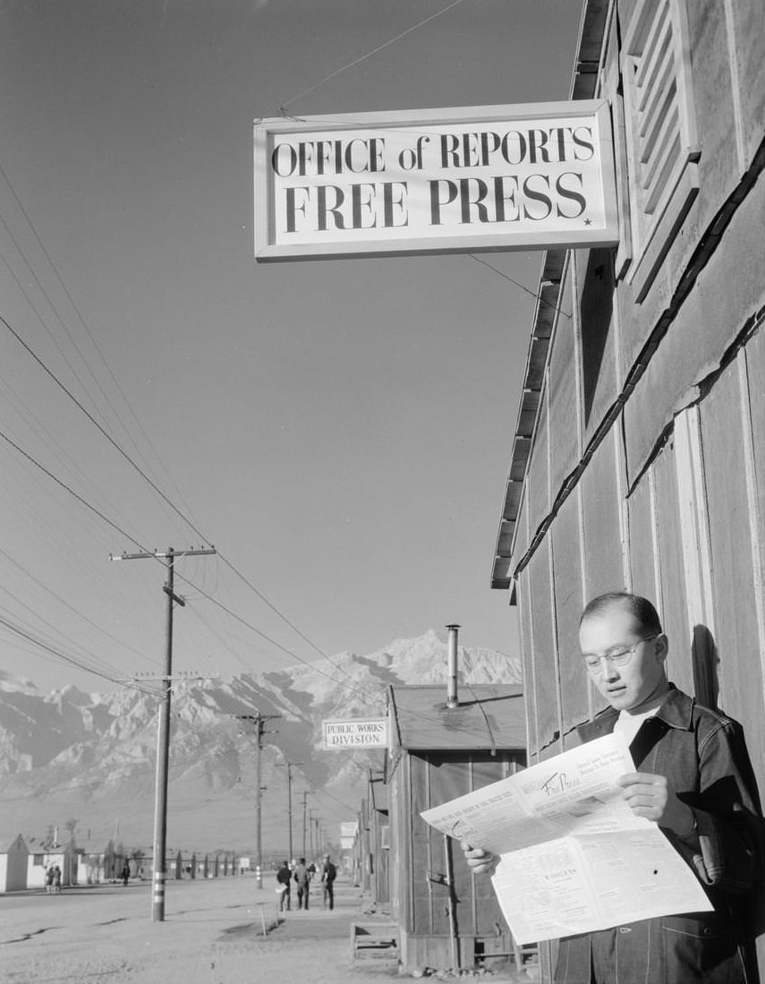 Photo shows editor Roy Takeno reading a copy of the Manzanar Free Press in front of the newspaper office at the Manzanar War Relocation Center; with mountains in the background.