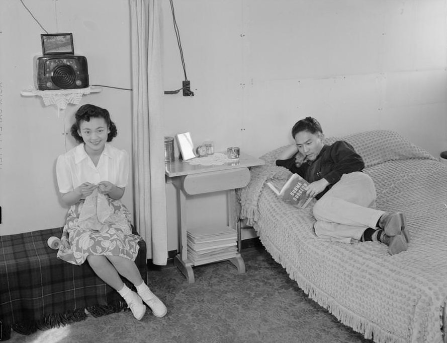 Mrs. Shimizu seated on couch with lacework, and Mr. Shimizu lying on bed, reading.